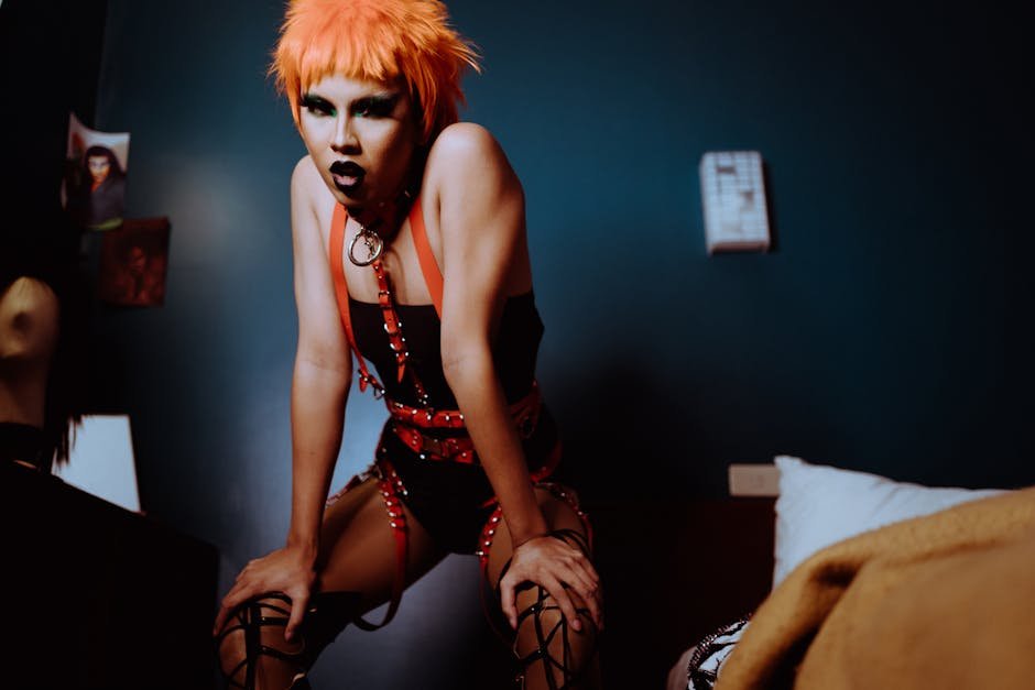 Qué es un Outfit? A confident drag queen with vibrant makeup and orange hair striking a pose indoors, exuding individuality and style.